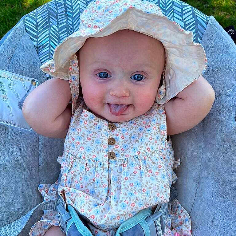 Billie is registered to the contest to win money with this photo: baby, child, infant, hat, sunhat, dress, floral, tongue_out, cute, smiling, outdoor, greenery, seat, cushion, playful, comfort, summer, sunshine, baby_accessory, relaxed