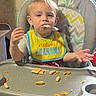 toddler, child, high_chair, bib, food, snack, spoon, messy, indoor, face, person, clothing, baby, cute, sitting, table, wall, door, red_sock, expression