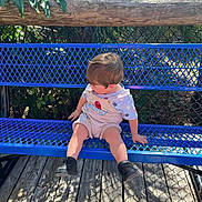 Zander is registered to the contest to win money with this photo: toddler, child, bench, blue_bench, outdoor, daylight, wooden_deck, greenery, plants, shorts, shirt, shoes, sitting, looking_down, metal_bench, fence, sunlight, shadow, playful, curious