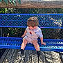 toddler, child, bench, blue_bench, outdoor, daylight, wooden_deck, greenery, plants, shorts, shirt, shoes, sitting, looking_down, metal_bench, fence, sunlight, shadow, playful, curious