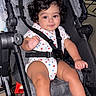 baby, stroller, child, infant, curly_hair, onesie, star_pattern, bottle, foot, hand, seatbelt, sitting, indoors, smile, cute, person, young_child, baby_clothes, baby_bottle, black_hair