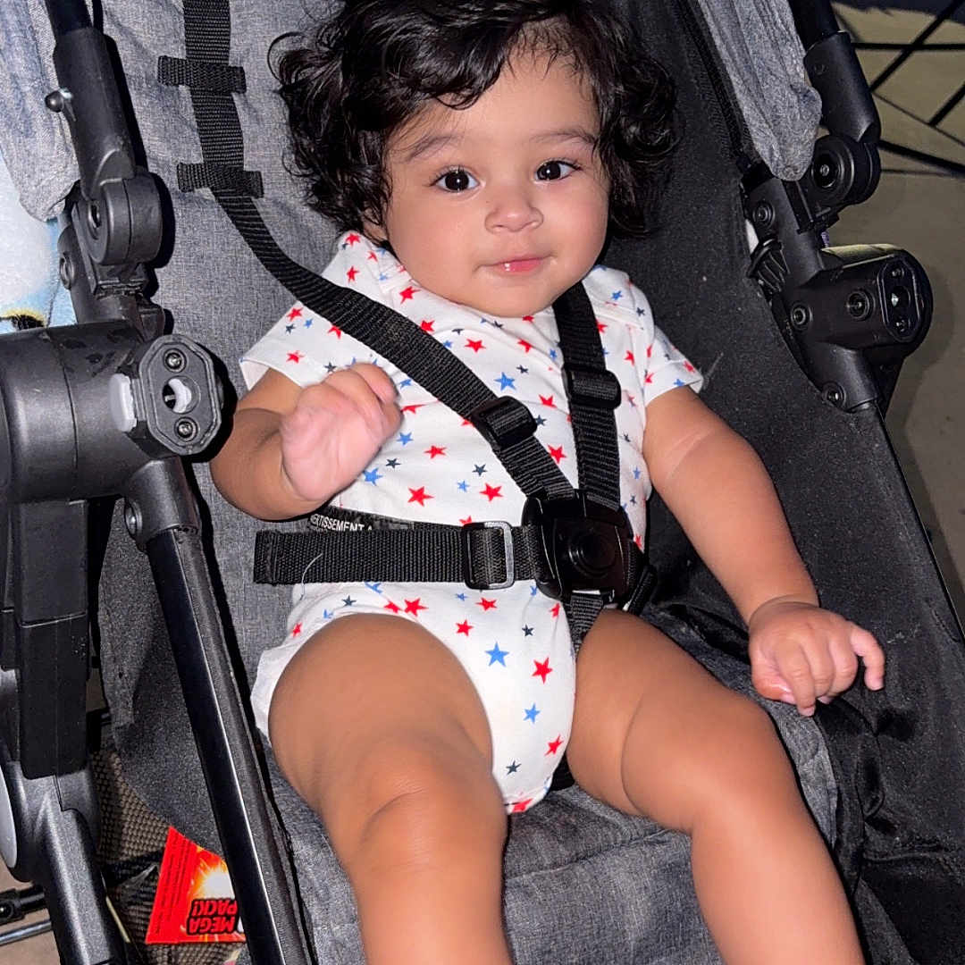 Andres is registered to the contest to win money with this photo: baby, stroller, child, infant, curly_hair, onesie, star_pattern, bottle, foot, hand, seatbelt, sitting, indoors, smile, cute, person, young_child, baby_clothes, baby_bottle, black_hair