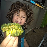 Juan Jose is registered to the contest to win money with this photo: child, broccoli, curly_hair, indoor, kitchen, food, vegetable, hand, blue_shirt, expression, face, person, carpet, countertop, bottle, cup, wall, curtain, toy, light