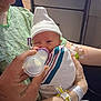 newborn, baby, infant, feeding, bottle, swaddle, hospital, hat, hand, tattoo, wristband, iv_line, medical_tag, striped_blanket, cabinet, person, portrait, sleepy, caregiver, nurse_area