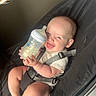 Vienna is registered to the contest to win money with this photo: baby, bottle, smiling, infant, baby_rocker, toy, hanging_toys, face, person, child, indoor, seat_belt, happy, milk, cute, legs, feet, clothing, bright, holding