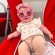 Vienna is registered to the contest to win money with this photo: baby, child, sunglasses, headband, red_chair, beige_pants, white_shirt, bunny_print, outdoor, relaxing, cute, face, person, infant, seated, summer, fashion, accessory, smiling, leisure