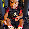 Zymir is registered to the contest to win money with this photo: baby, child, infant, car_seat, sitting, portrait, face, eyes, smiling, clothing, hat, sneakers, sportswear, basketball, black_and_red, indoor, vehicle, harness, fashion, cute