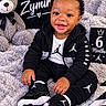 Zymir is registered to the contest to win money with this photo: baby, infant, child, smiling, portrait, sitting, indoor, teddy_bear, blanket, pillow, black_clothing, sportswear, sneakers, jordan_brand, milestone_photo, six_months, curly_hair, happy, cozy, close_up