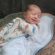 Asher is registered to the contest to win money with this photo: arm, baby, blanket, child, cozy, cute, dinosaur_pattern, face, indoors, infant, newborn, onesie, peaceful, person, resting, sleeping, sleepwear, smiling, soft_fabric, tattoo