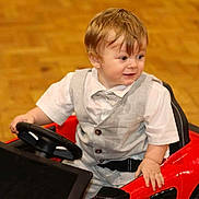 Oskar joined the competition — help win amazing prizes! toddler, child, toy_car, steering_wheel, vest, bow_tie, white_shirt, indoor, wooden_floor, red, smiling, person, cute, seatbelt, short_hair, play, happy, young_child, car, floor