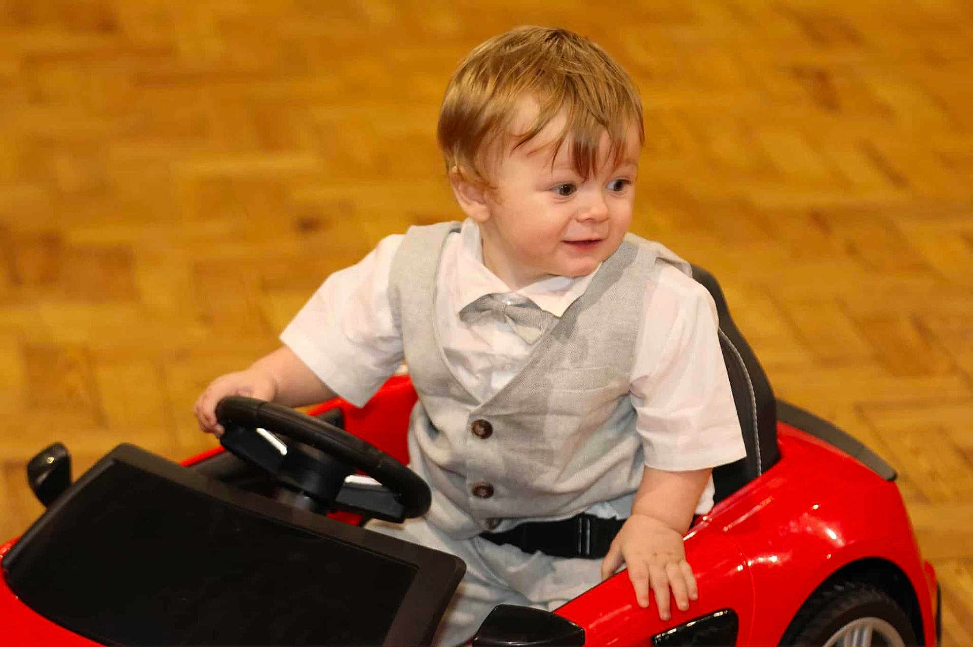 Oskar joined the competition — help win amazing prizes! toddler, child, toy_car, steering_wheel, vest, bow_tie, white_shirt, indoor, wooden_floor, red, smiling, person, cute, seatbelt, short_hair, play, happy, young_child, car, floor