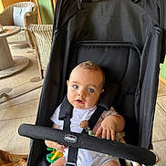 Andréa a rejoint le concours — aidez-le/la à gagner de superbes lots ! baby, stroller, child, blue_eyes, toy, giraffe, black_stroller, outdoor, patio, wicker_chair, table, floor, clothing, infant, person, seatbelt, curious, cute, sitting, daylight
