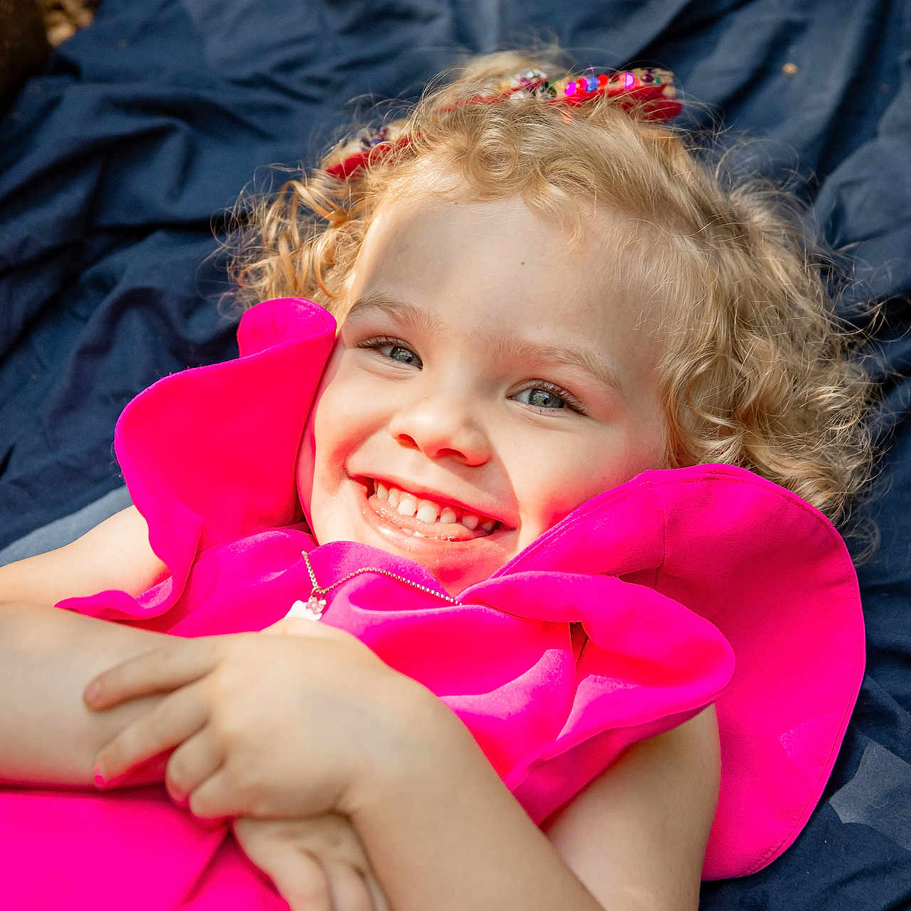 Kelly is registered to the contest to win money with this photo: accessory, blanket, blonde, child, childhood, curly_hair, cute, face, fashion, girl, happy, joyful, lying_down, outdoor, person, pink_dress, playful, portrait, smiling, sunlight