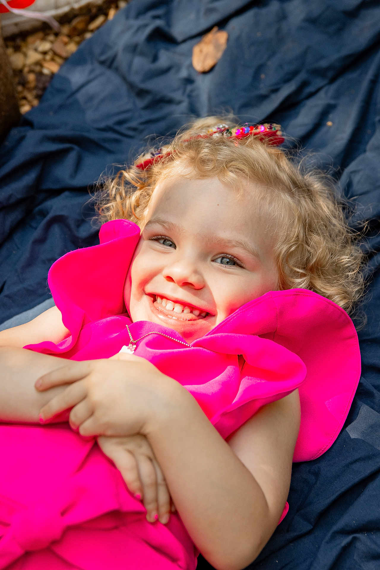Kelly is registered to the contest to win money with this photo: child, girl, smiling, curly_hair, blonde, pink_dress, outdoor, blanket, happy, face, portrait, cute, joyful, childhood, fashion, person, lying_down, playful, sunlight, accessory