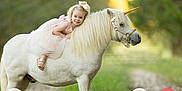 Kelly is registered to the contest to win money with this photo: animal, blonde, child, curly_hair, cute, dress, girl, grass, happy, horn, nature, outdoor, path, pet, photography, pink_dress, pony, riding, smile, unicorn