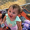 baby, child, smiling, outdoor, chain_link_fence, blanket, toy, purple_toy, grass, sunlight, happy, person, clothing, play, hand, sitting, pants, top, face, expression