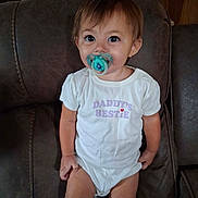 Michaela is registered to the contest to win money with this photo: toddler, child, pacifier, onesie, white_clothing, sitting, cushioned_chair, brown_furniture, indoor, curious_expression, short_hair, baby, young_child, furniture, wooden_cabinet, home, portrait, casual, cute, small_child
