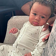 Cordell is registered to the contest to win money with this photo: baby, child, infant, onesie, stars, couch, hand, curly_hair, portrait, indoor, cozy, furniture, person, cute, comfortable, skin, face, expression, seated, support