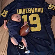 Lyonel is registered to the contest to win money with this photo: baby, infant, football, sports_jersey, clothing, blue, number_19, underwood, lying_down, small_football, baby_clothes, sock_shoes, bed, blanket, person, newborn, cute, sports_theme, indoor, resting