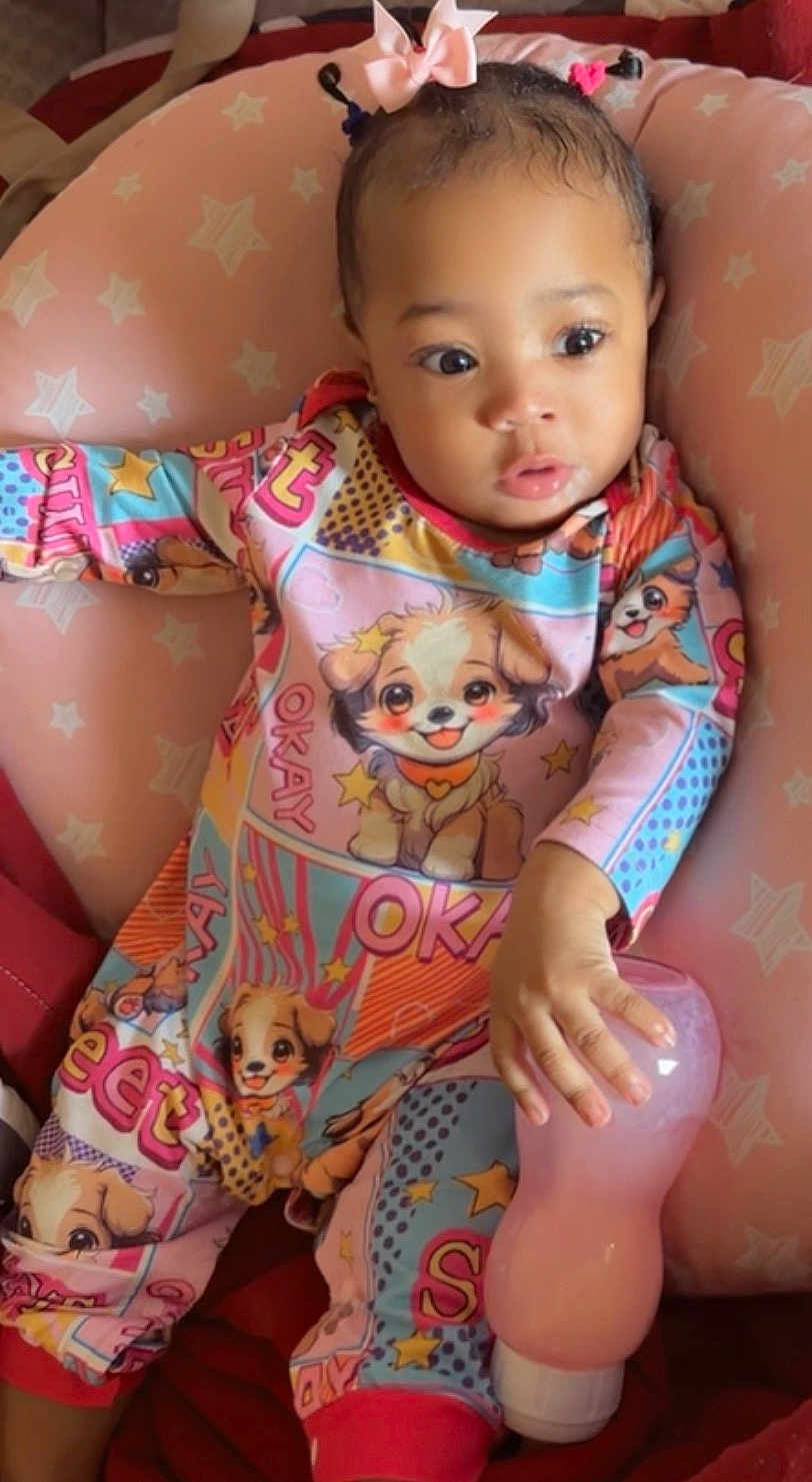 Kloie is registered to the contest to win money with this photo: baby, infant, child, pajamas, onesie, bottle, feeding_bottle, pink, bow, headband, baby_seat, cushion, stars, cartoon_dog, hand, big_eyes, portrait, cute, curious_expression, seated