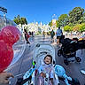 amusement_park, baby, balloon, blue_sky, child, crowd, family, fan, infant, outdoor, park, portrait, ride, seated, smiling, stroller, sunny, toy, travel, trees