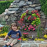 child, smiling, outdoor, flower_bed, stone_wall, greenery, plant, cup, shoes, shorts, tshirt, garden, nature, sunlight, happy, portrait, casual, young_child, colorful, daytime
