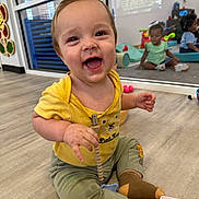 Kasen joined the competition — help win amazing prizes! toddler, baby, child, smiling, sitting, yellow_shirt, green_pants, pacifier_clip, mirror, reflection, playroom, floor, toy, indoor, happy, cute, childcare, infant, casual_clothing, playful