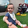 baby, child, grass, toy_truck, basketball, bib, stroller, outdoor, smiling, sitting, short_sleeve, sock, window, building_wall, toy, playtime, sunlight, face, person, daytime