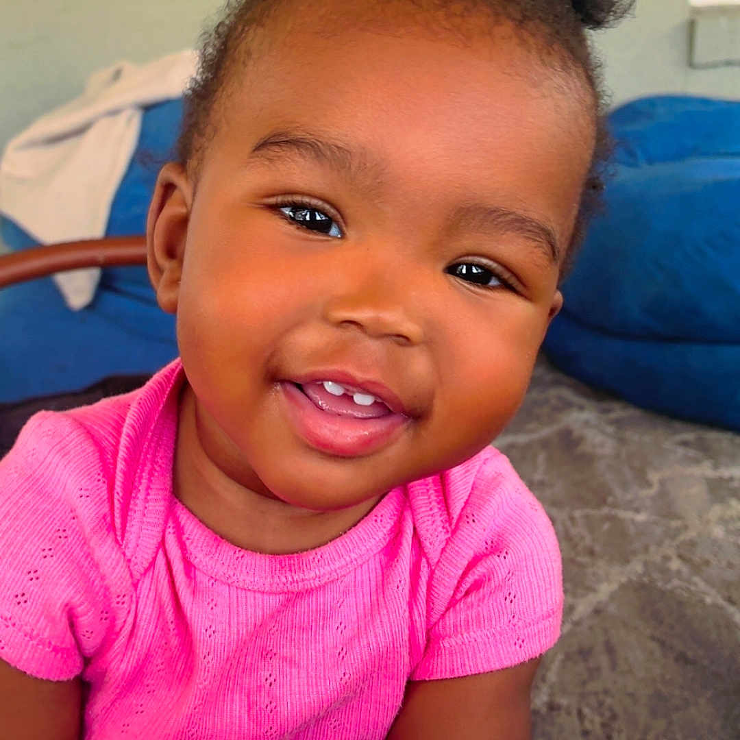 Jaziriah joined the competition — help win amazing prizes! toddler, child, smiling, baby, pink_shirt, hair_puffs, hand, holding_hands, indoor, carpet, sofa, portrait, cute, happy, person, skin, face, young_child, seated, closeup