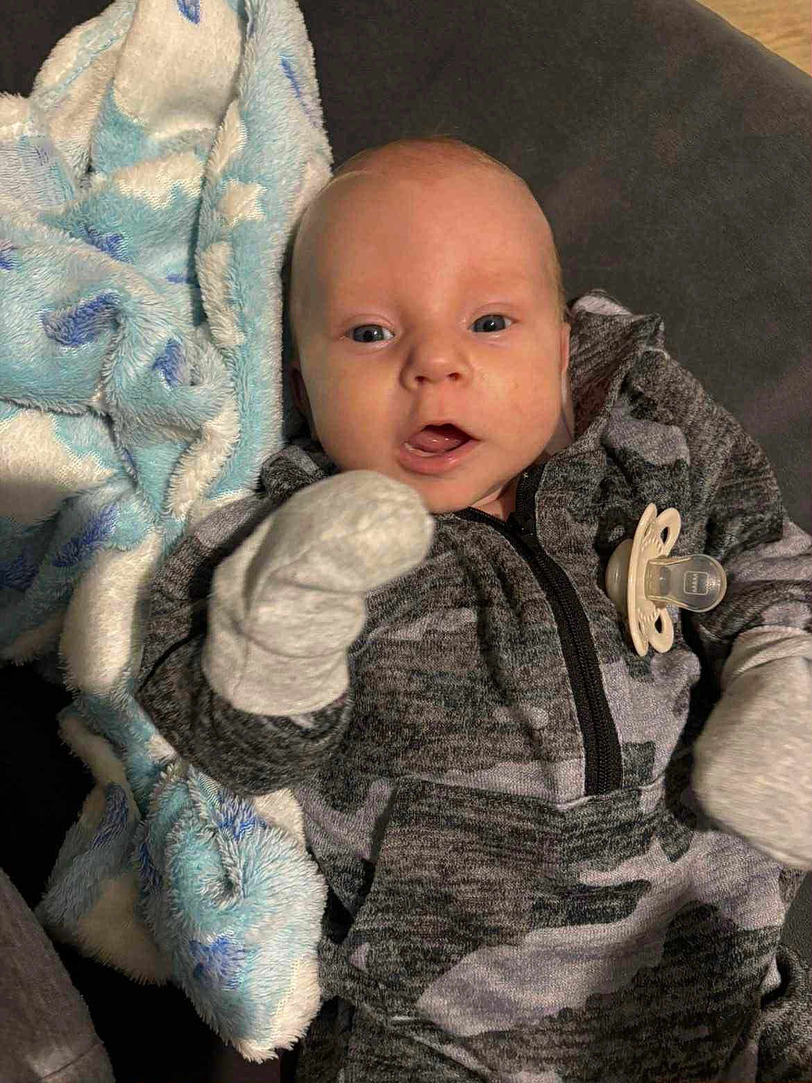 Layton is registered to the contest to win money with this photo: baby, infant, blanket, mittens, pacifier, clothing, face, person, cute, portrait, lying_down, blue, gray, soft_texture, young_child, indoor, expression, cozy, closeup, newborn