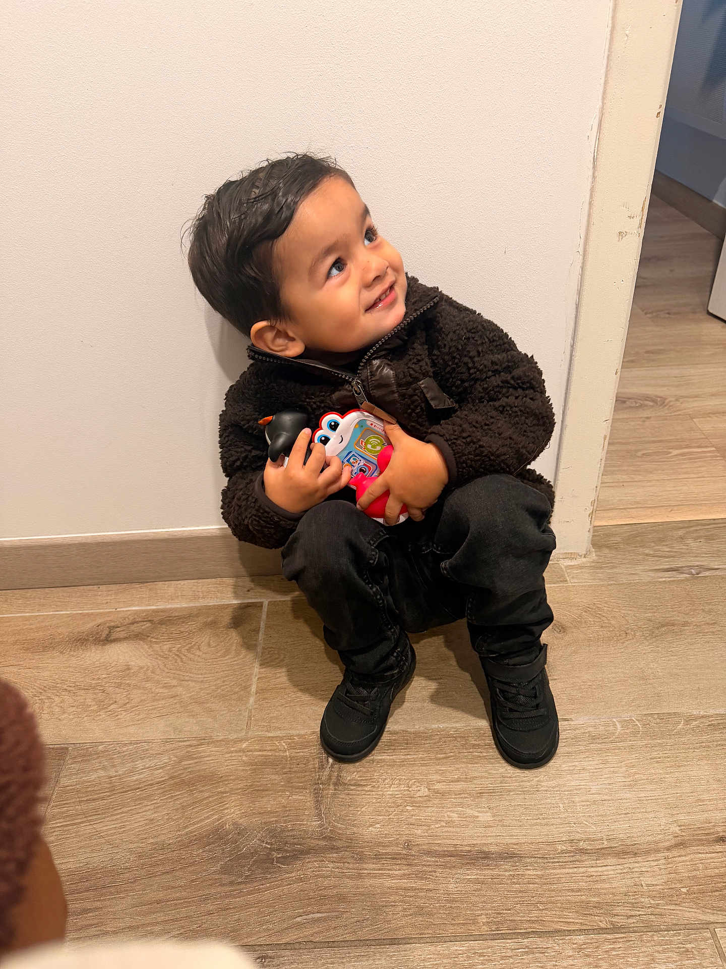 Lyano a rejoint le concours — aidez-le/la à gagner de superbes lots ! child, toddler, smiling, toy, indoor, hardwood_floor, white_wall, sitting, jacket, black_shoes, looking_up, playful, face, hair, eyes, door_frame, candid, casual_clothing, small_handheld_toy, floor_tile
