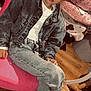 child, denim_jacket, jeans, sneakers, pink_chair, indoor, wooden_floor, blanket, floral_pattern, face, person, seated, crossed_legs, casual_clothing, young, curious_expression, hair, fashion, cozy, furniture