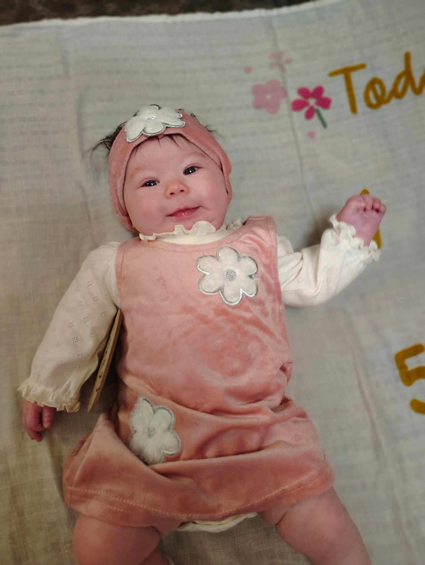 Blakely is registered to the contest to win money with this photo: baby, infant, smiling, pink_dress, headband, flower_applique, blanket, lying_down, portrait, cute, hand, legs, eyes, mouth, soft_fabric, bedroom, skin, clothing, bokeh_background, peaceful