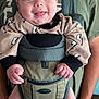 Arlus joined the competition — help win amazing prizes! baby, infant, smiling, baby_carrier, person, clothing, beige_outfit, lightning_bolt_pattern, hands, indoor, adult, green_shirt, babywearing, cute, face, tongue, happy, closeup, portrait, holding