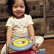 Alisson Abril joined the competition — help win amazing prizes! child, toddler, smile, toy_car, steering_wheel, bow, curly_hair, pajamas, sitting, wooden_dresser, drawer_knobs, hardwood_floor, indoor, portrait, play, hands, feet, happy, camera, cute