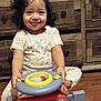 child, toddler, smile, toy_car, steering_wheel, bow, curly_hair, pajamas, sitting, wooden_dresser, drawer_knobs, hardwood_floor, indoor, portrait, play, hands, feet, happy, camera, cute