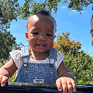 Anaya is registered to the contest to win money with this photo: toddler, child, smiling, denim_overalls, beaded_necklace, outdoor, sunlight, trees, blue_sky, greenery, hands, portrait, baby, happy, person, nature, daylight, cute, clothing, face