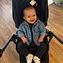 baby, smile, denim_jacket, black_outfit, stroller, wooden_floor, indoor, child, footwear, white_shoes, happy, sitting, cute, infant, person, casual_clothing, small_child, baby_shoes, cheerful, portrait