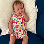 baby, infant, child, couch, pillow, romper, colorful_clothing, fruit_pattern, hand, thumb_sucking, hair, face, legs, barefoot, indoors, seated, expression, cute, comfortable, blue_couch