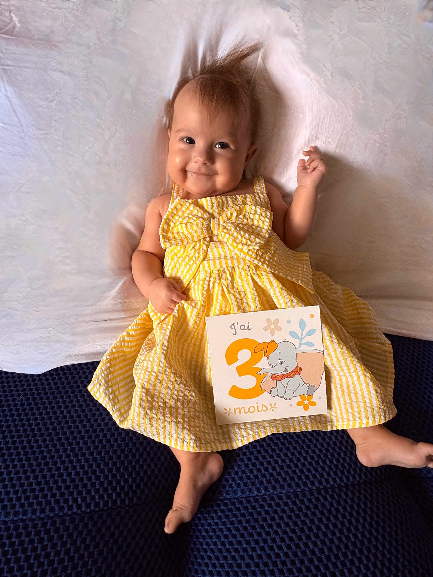 Alba a rejoint le concours — aidez-le/la à gagner de superbes lots ! baby, infant, yellow_dress, bow, milestone_card, elephant_illustration, smile, lying_down, blue_surface, white_background, cute, happy, child, portrait, indoors, clothing, skin, hair, hand, foot