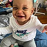 baby, smiling, sitting, indoor, clothing, pacifier, blanket, face, person, happy, headwear, toy, nike, pants, floor, fan, room, hair, child, expression