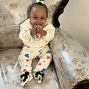 Annalise is registered to the contest to win money with this photo: baby, child, smiling, sitting, toy, mickey_mouse, clothing, couch, vintage_furniture, floral_pattern, footwear, hair_bow, indoors, cute, happy, person, infant, playful, comfort, apparel