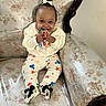 baby, child, smiling, sitting, toy, mickey_mouse, clothing, couch, vintage_furniture, floral_pattern, footwear, hair_bow, indoors, cute, happy, person, infant, playful, comfort, apparel