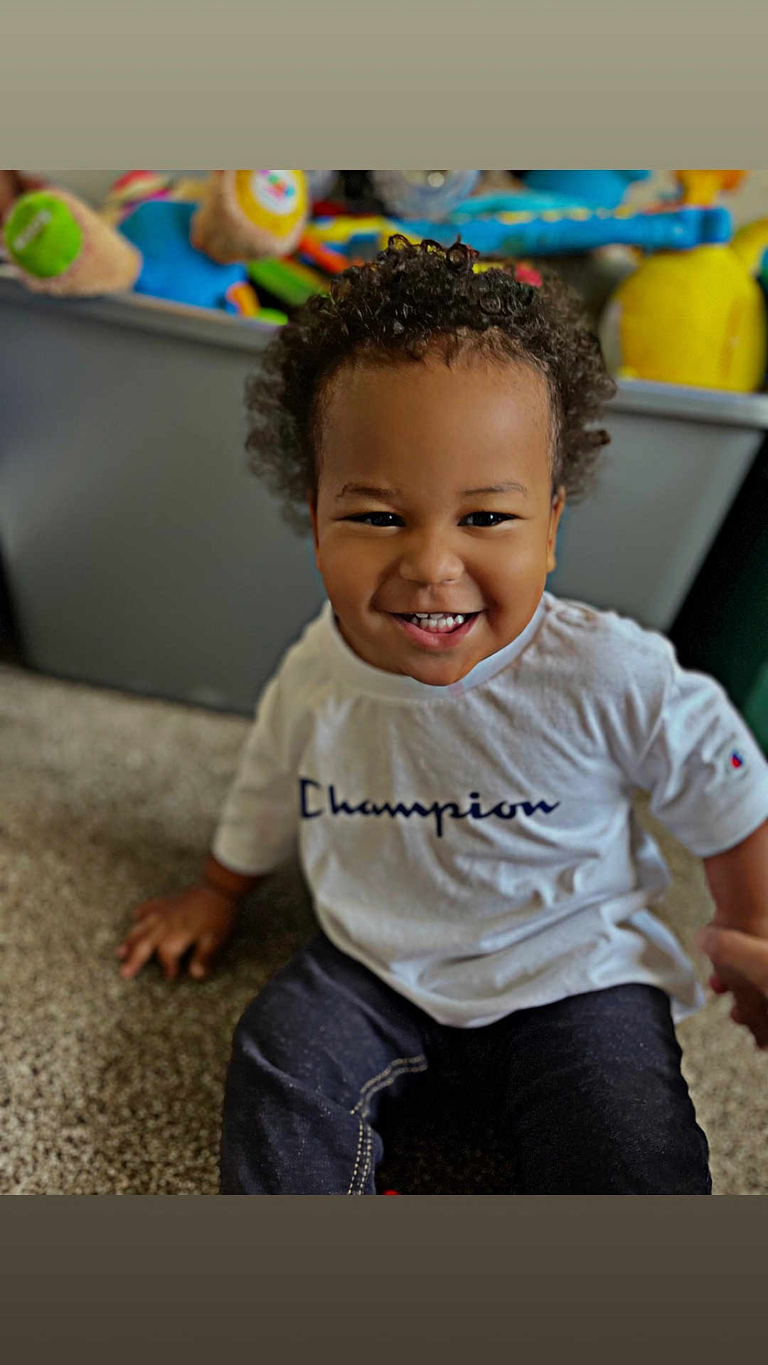 Zaidan joined the competition — help win amazing prizes! toddler, child, smiling, curly_hair, white_shirt, champion_logo, carpet, toys, indoor, happy, person, sitting, face, clothing, casual, young_child, playroom, background_blur, cute, portrait
