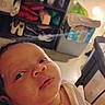 baby, newborn, infant, face, portrait, indoor, home, lying_down, looking_at_camera, eyes, nose, mouth, skin, clothing, cream_outfit, shelves, clothes, lamp, stroller, close_up