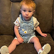 Brooklyn is registered to the contest to win money with this photo: baby, brown_chair, casual, child, clothing, cute, denim_overalls, expression, face, footwear, indoor, looking_up, person, portrait, seat, short_hair, sitting, toddler, white_sandals, young_child