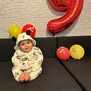 Asser a rejoint le concours — aidez-le/la à gagner de superbes lots ! baby, child, mickey_mouse, balloon, number_balloon, red_balloon, yellow_balloon, couch, black_couch, hooded_outfit, smiling, sitting, indoor, celebration, birthday, cute, wall, patterned_wall, foot, happy