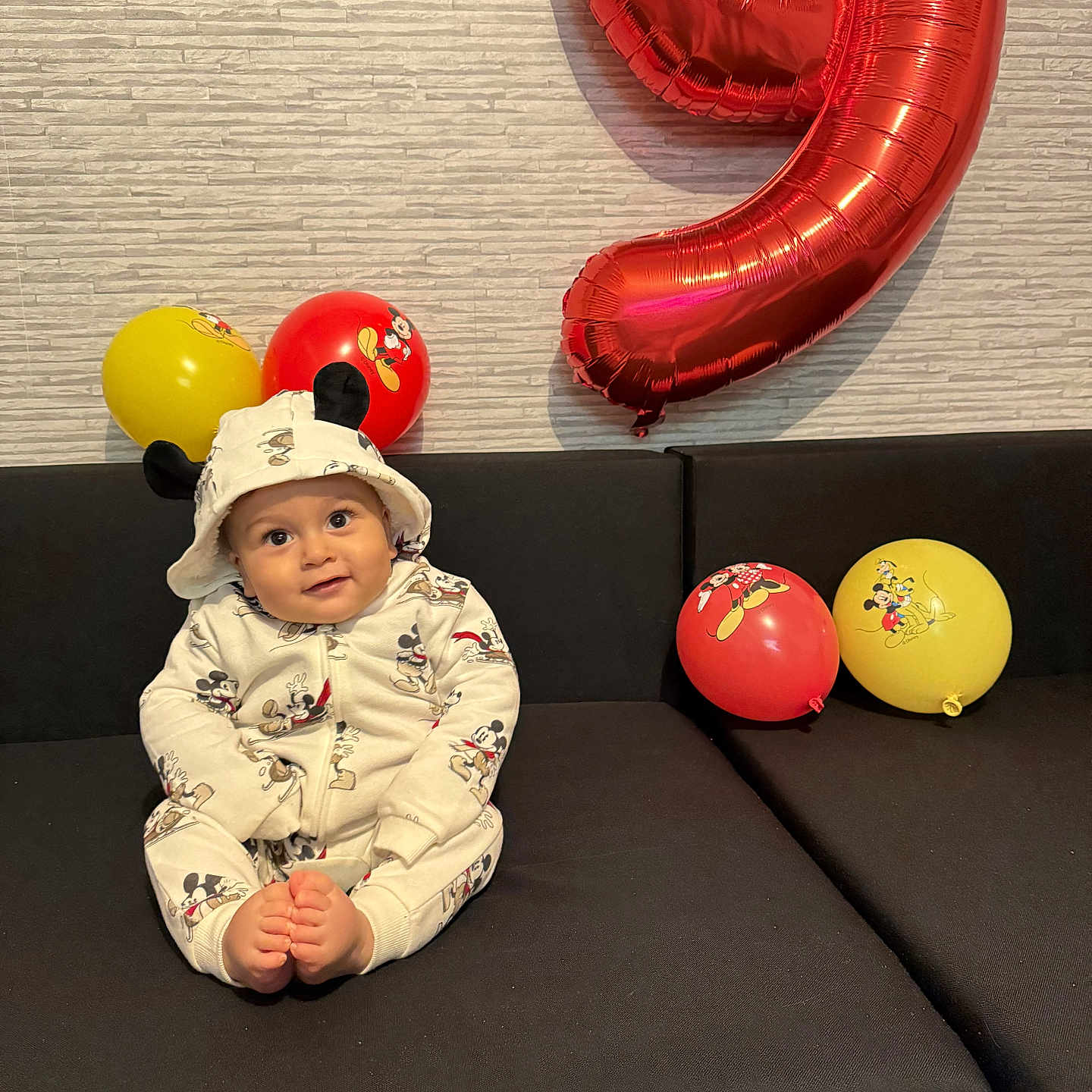 Asser a rejoint le concours — aidez-le/la à gagner de superbes lots ! baby, balloon, birthday, black_couch, celebration, child, couch, cute, foot, happy, hooded_outfit, indoor, mickey_mouse, number_balloon, patterned_wall, red_balloon, sitting, smiling, wall, yellow_balloon
