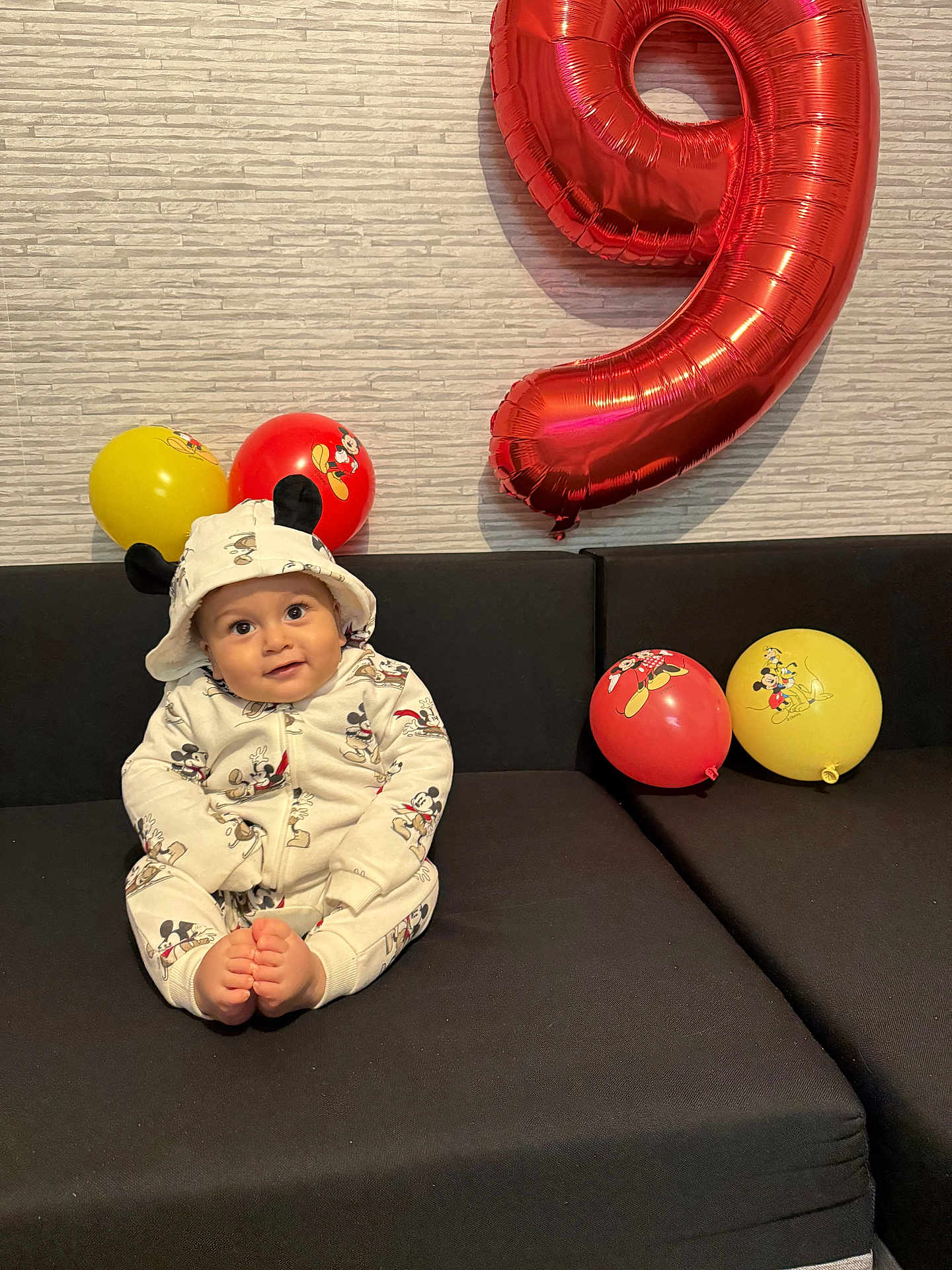 Asser a rejoint le concours — aidez-le/la à gagner de superbes lots ! baby, child, mickey_mouse, balloon, number_balloon, red_balloon, yellow_balloon, couch, black_couch, hooded_outfit, smiling, sitting, indoor, celebration, birthday, cute, wall, patterned_wall, foot, happy