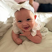 Nellie is registered to the contest to win money with this photo: baby, headband, smiling, face, child, bed, white_clothing, infant, cute, happy, portrait, closeup, soft_lighting, indoors, person, skin, chubby_cheeks, hand, lying_down, background_blur